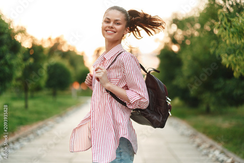 Fotografie Young woman with striped shirt and backpack walks along a sunny park path, smiling confidently as she glances back and enjoys a carefree moment