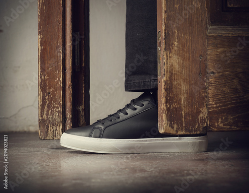 Close-up Action Shot of Foot in Black Sneaker with White Sole in Doorway of Room with Wooden Frame and Concrete Floor from Low Viewpoint