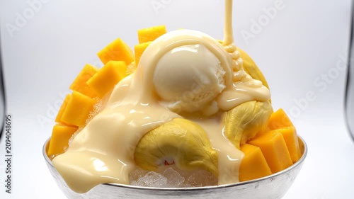 Sweet condensed milk pouring over a bowl of shaved ice dessert with fresh mango cubes and durian fruit, studio lighting.