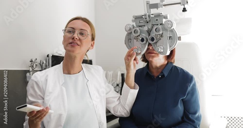 Professional ophthalmologist testing eyesight of middle aged woman patient in medical clinic using phoropter adjusting modern apparatus asking questions about vision clarity before prescribing glasses