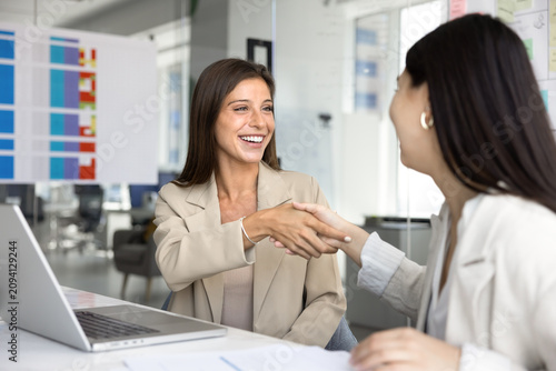 Foto Two professional women smiling and shaking hands seated at desk with laptop and papers in modern office, gesture of mutual respect, successful negotiation outcome, and perspective collaboration