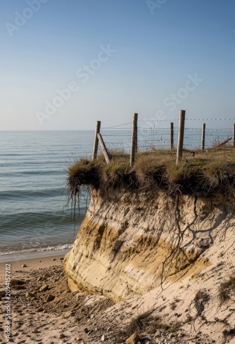 Coastal erosion of a sandy bluff with a weathered fence and exposed roots against a clear blue sky for climate change and environmental stories