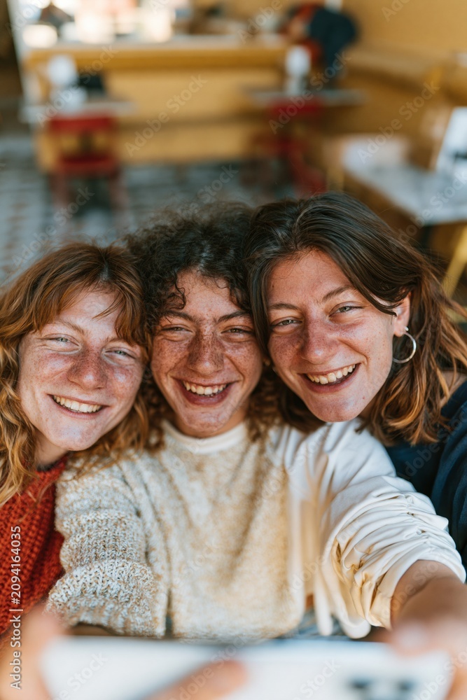Fototapeta premium Three joyful young women with freckled skin smiling for a close-up selfie in a cafe for social media and friendship lifestyle concepts