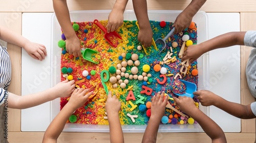 Children's hands engaging in sensory play with a vibrant bin of colorful rice, beads, and small toys, fostering tactile exploration and learning.