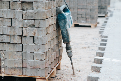 A construction site features stacks of gray bricks arranged neatly on wooden pallets. A drill is positioned against a pile of bricks, ready for use in the building process