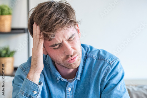 Obraz na płótnie Young man holding his head in pain sitting on a couch, stressed and suffering fr