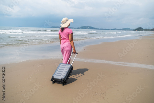 Young woman in pink dress and straw hat strolling leisurely on a sandy beach pulling a grey suitcase with sea and cloudy sky in background. Leisure arrival concept