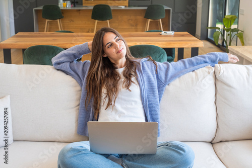 Young woman relaxing on a sofa with a laptop, stretching and smiling while working from home in a bright modern living room, feeling comfortable and content.