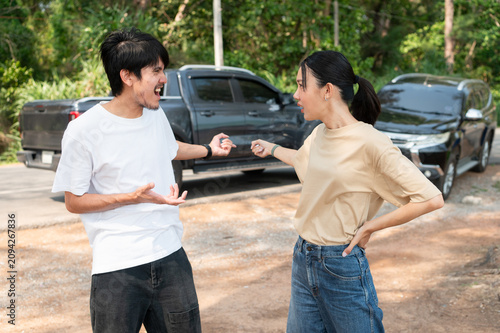 Two people involved in a roadside dispute argue beside their cars, showing shock and frustration after a sudden accident.