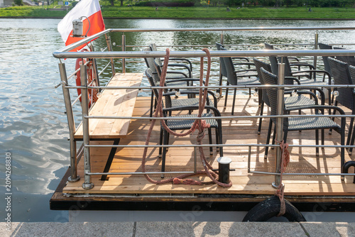 Принти на полотні Empty open-air passenger deck with rows of black woven chairs, wooden flooring, metal railings, mooring ropes and a Polish flag on a vessel docked on the Vistula River in Krakow