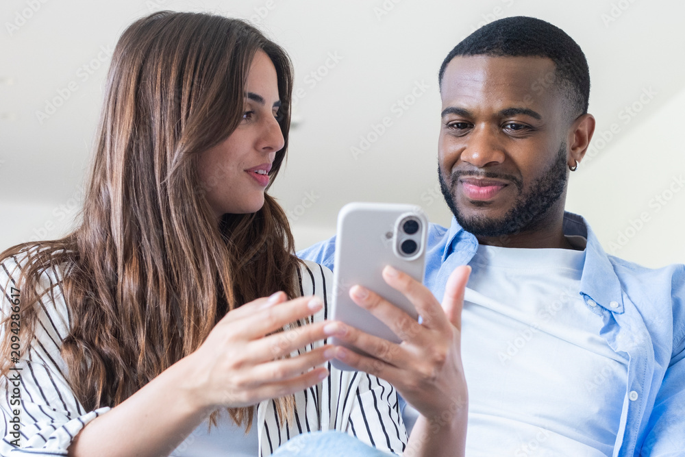 Fototapeta premium Young couple looking at smartphone together at home, smiling and sharing a moment of connection, technology, relationship and casual lifestyle