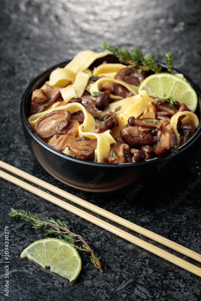Fototapeta premium Shimeji and shiitake mushrooms stir-fried with soy sauce, garlic and thyme, served with noodles and lime slice in a black ceramic bowl.