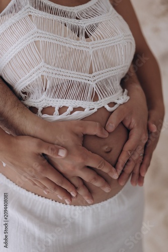 Close-up view of couple hands forming heart shape on pregnant belly with white macrame top