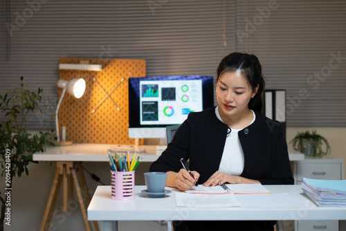An Asian businesswoman sits at her office desk at night, focused on writing in a notebook.