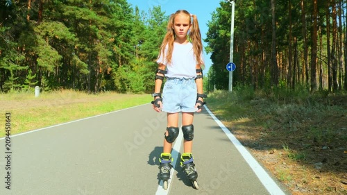 Child girl in casual clothes roller skating on road in forest on sunny summer day.
