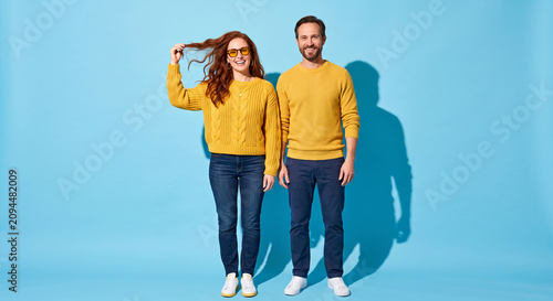Full length studio portrait of smiling young couple wearing yellow sweaters and blue jeans standing together against a light blue background