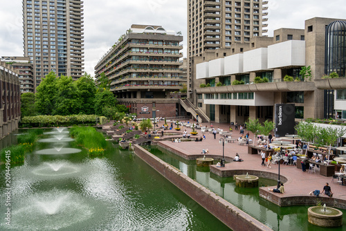 Canvas Print The bustling lakeside terrace of the Barbican Estate in London, UK, featuring fountains spouting water in a green-tinted canal in the foreground, with with people relaxing on the brick plaza