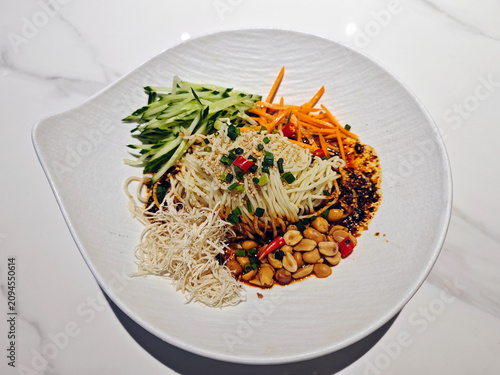A delicious plate of Chinese cold noodles topped with shredded chicken, fresh cucumber, carrots, crunchy peanuts, and spicy chili oil