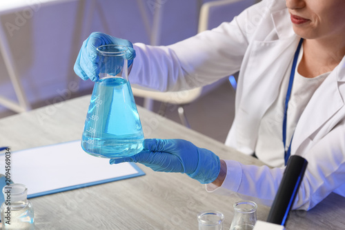 Scientist in lab coat and blue gloves holding an erlenmeyer flask with blue l...
