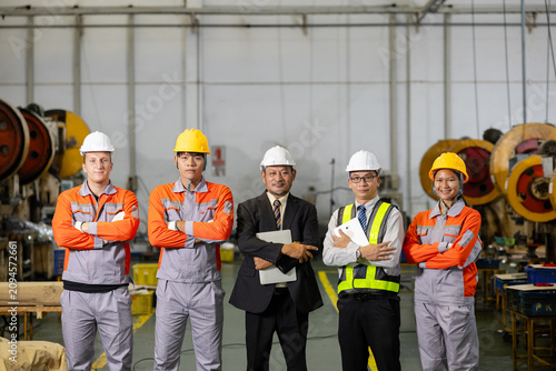 Diverse team of engineers and factory workers in safety gear posing with manager in an industrial workshop, wearing hard hats and uniforms, showcasing teamwork and manufacturing.