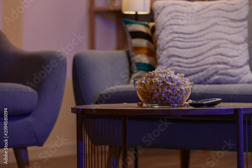 Tasty popcorn with TV remote controller on table in living room at night