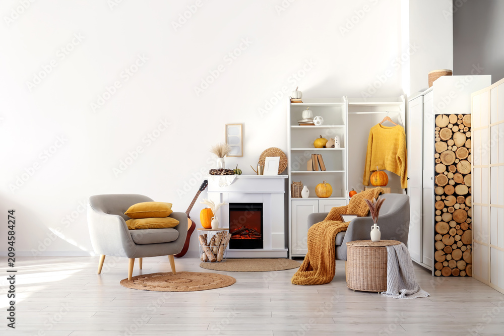 Fototapeta premium Interior of living room with armchairs, coffee tables, guitar, autumn home decor on fireplace and pumpkins in shelving unit near white wall