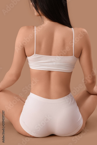 Картината върху платно Beautiful young woman in white underwear sitting on brown background, back view