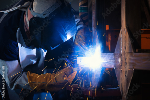 Welder in protective gear works closely on metal. Bright sparks fly from the welding torch. The worker's posture shows deep concentration. Industrial workshop environment features heavy machinery