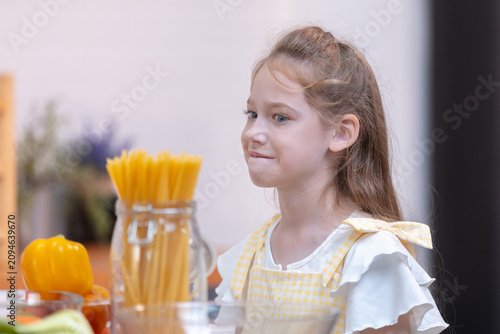 Fotografie Young Caucasian girl standing in home kitchen looking at dry spaghetti pasta wit
