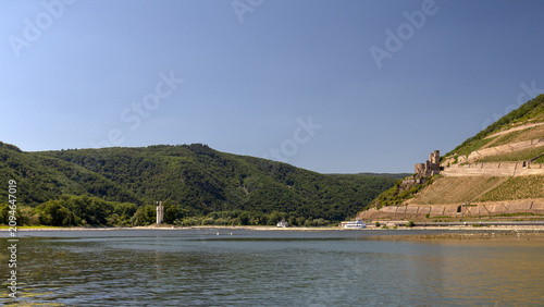 Wide scenic view of the Rhine River near Bingen, Germany, featuring the historic Mouse Tower, Ehrenfels Castle ruins, steep vineyards, forested hills and passenger boats on a sunny summer day.
