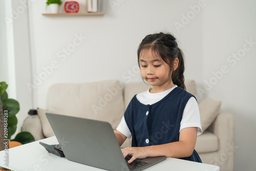 Young girl sitting at white table using laptop computer in living room environment with focused and engaged expression, casual indoor activity for children learning