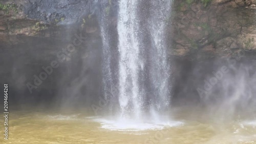 Wide horizontal shot of a scenic waterfall crashing into a muddy river below, generating thick mist and water spray.