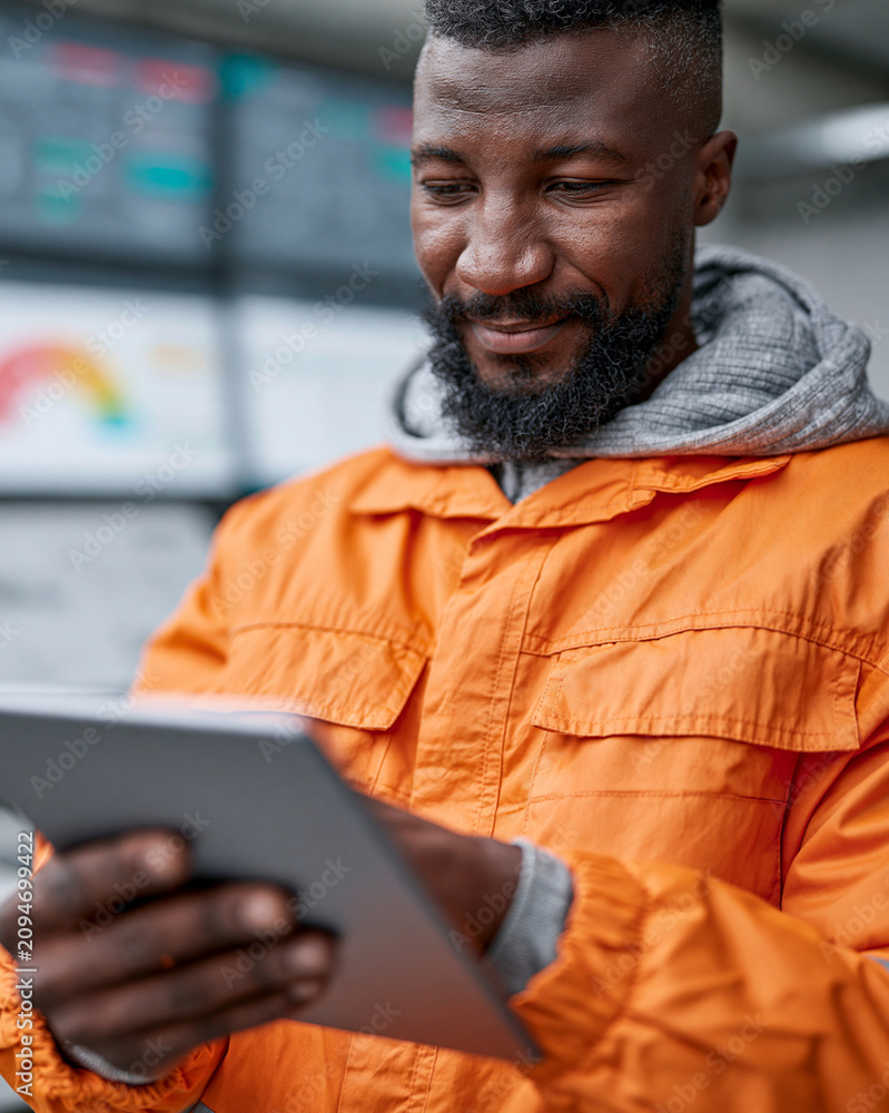 Fototapeta premium Man smiles while using a tablet. He wears an orange jacket and grey hoodie. His eyes show satisfaction and focus. He stands near a data screen display. Ideal for tech, business