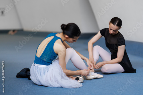Two ballet dancers tying pointe shoes during rehearsal in a studio, preparing for performance and practicing technique and teamwork