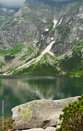 Clear lake reflects steep rocky mountains with snow patches. Beautiful alpine pond stands surrounded by green pine trees and rocks. Deep water of pristine lake creates tranquil atmosphere. vertical
