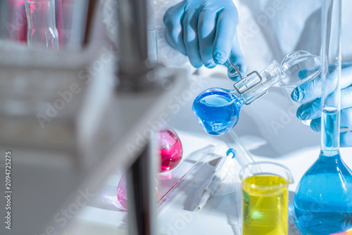Chemistry laboratory scene with colorful chemical solutions in glass flasks and cylinders, scientist in gloves writing results, analytical experiment workspace.