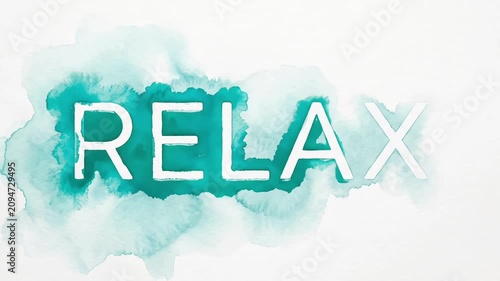 The word RELAX cutout from white paper, revealed by expanding teal watercolor paint spreading on textured paper