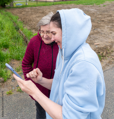 Принти на полотні A smiling younger woman in a blue hoodie shows smartphone content to an elderly woman wearing glasses during an outdoor walk