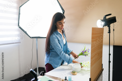 Young female photographer arranging prop flowers and a cosmetic product for a creative flat lay still life setup
