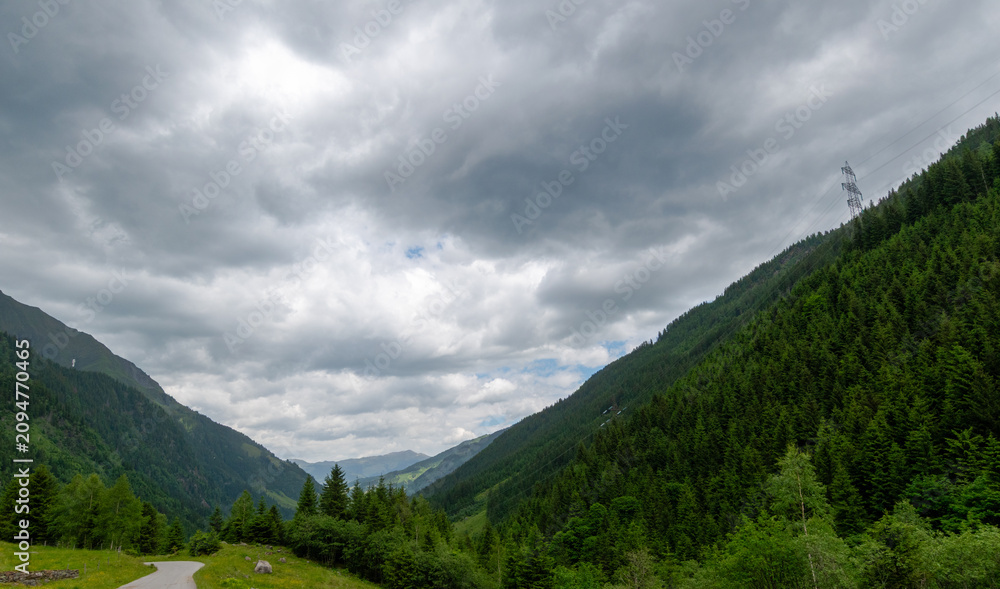 Fototapeta premium Winding road through a forested alpine valley under storm clouds