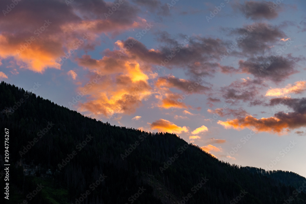 Obraz premium Dramatic alpine sunset cloudscape over silhouetted mountain ridges and pine forest, featuring glowing orange clouds against a deep blue sky, Dolomites, South Tyrol, Italy