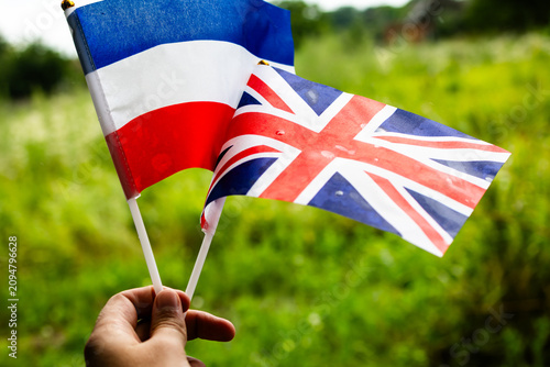 Hand holds small flags of France and the United Kingdom waving together in a lush green park symbolizing friendship bilingual culture travel and diplomacy under bright summer light