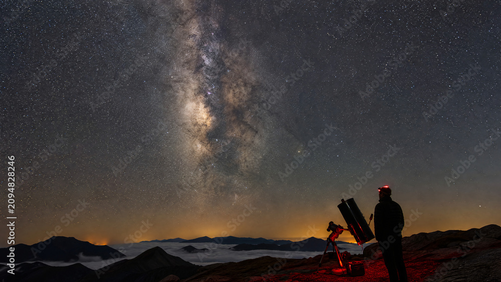 Fototapeta premium Amateur astronomer with telescope and red headlamp watching the Milky Way above a sea of clouds