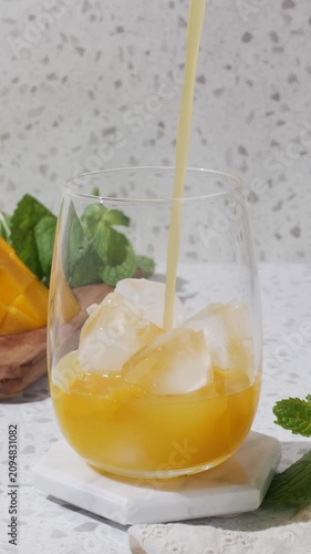 Summer mango drink mocktail with ice and mint on a white coaster, styled in bright sunlight on a terrazzo countertop with fresh mango and clean copy space. A clear rounded glass holds a golden mango
