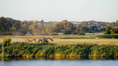 Photos A tractor rakes mown grass in a meadow by the river, against a backdrop of trees