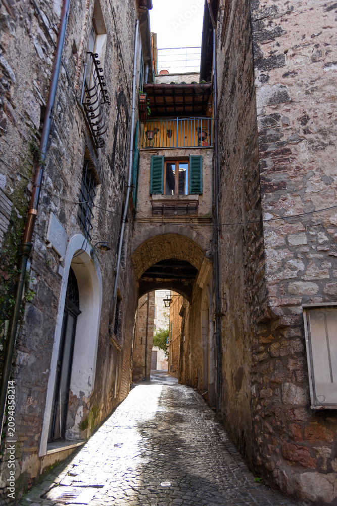 Fototapeta premium Vertical perspective of a narrow cobblestone street passing under a historic stone archway with traditional shuttered windows and balconies in the medieval hill town of Stroncone, Umbria, Italy