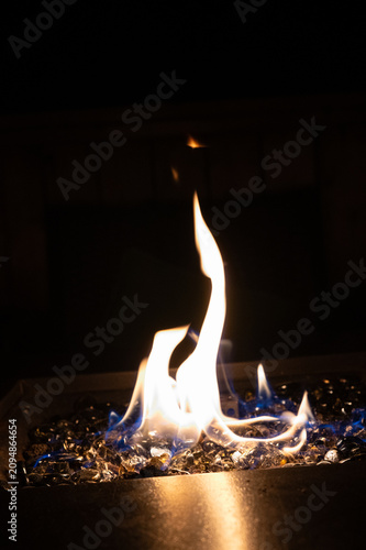 A close-up of a propane-fueled fire pit with bright orange flames and a blue base flickering over dark, crystalline fire glass at night