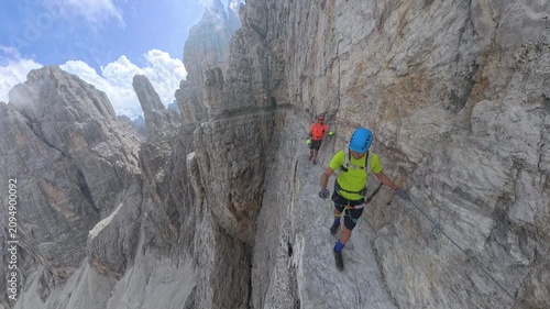 Two hikers crossing an exposed ledge on Via Ferrata Bocchette Centrali with dramatic drops below, Brenta Dolomites