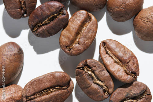 Coffee Beans Displayed In Tight Close Up Showing Roasted Texture And Surface Cracks On White Background With Directional Lighting And Distinct Cast Shadows.