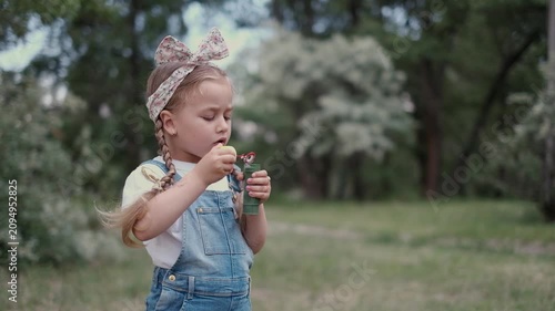 Cute little girl blowing soap bubbles in summer park with boy playing in background